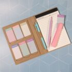 A weekly planner and notepad on a blue geometric surface. The planner has sticky notes labeled with days of the week, and two pastel pens, pink and purple, lie on the open notepad beside a pink highlighter.