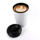 A white travel coffee mug with a black lid next to it is filled with coffee, featuring intricate latte art in the shape of a heart and leaves on the foam.