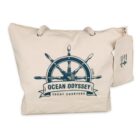 A cream-colored tote bag featuring a graphic of a ships wheel with the words Ocean Odyssey Yacht Charters printed on it. The bag has a small attached pouch with an anchor design.