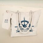 Canvas tote bag on sand, featuring a nautical design with a ships wheel and rope handles. An attached tag with an anchor graphic and text hangs from the strap. The ocean is blurred in the background.