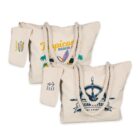 Two beige canvas tote bags with white rope handles. One bag features a tropical beach design with palm trees; the other shows a ships wheel with an anchor. Both have attached pouches with matching graphics.