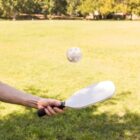 A person playing pickleball in a park, holding a paddle and hitting a perforated ball. The background features a lush green field and trees under a sunny sky.