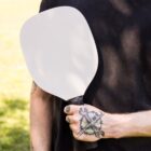 A person holding a pickleball paddle with a blank face and black handle, showing a tattoo of a wrench and hammer crossed on their hand. They are wearing a black shirt, and the background is a grassy area.