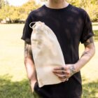 A person with tattoos, wearing a black t-shirt, holds a beige drawstring bag while standing on a grassy field. Trees are visible in the background under a clear sky.
