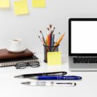 A tidy workspace with a laptop, a cup of coffee, a stack of books, glasses, pens, brushes in a holder, and sticky notes on the wall. A pen is placed prominently in front of the laptop.