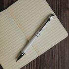 A silver pen rests on an open, lined notebook with a beige cover. The notebook is placed on a dark wooden surface, creating a contrast between the light pages and the rich wood grain.