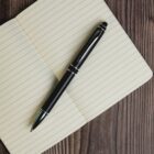 A black pen lies diagonally on an open, lined notebook placed on a wooden surface.