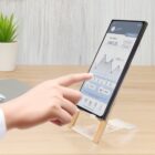 A person touches a smartphone on a wooden stand, displaying a finance app with a graph showing a budget of $567.35. A potted plant and a closed laptop are in the background on a wooden surface.