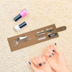 A nail care kit with scissors, clippers, a file, and other tools lies open on a beige textured surface. Nearby are pink and white nail polish bottles. Two hands display blue and patterned manicured nails.