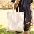 A person stands outdoors holding a plain beige tote bag. They are wearing a black shirt and camo shorts. Tattoos are visible on their arm. A grassy park area and trees are in the background under a clear sky.