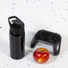 A black water bottle, a video game controller, and a red apple are placed on a white speckled surface.