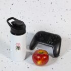 A black game controller, a red apple, and a white water bottle with a black lid and Koala Kuddles graphic are placed on a speckled white surface.