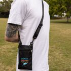 A person wearing a white T-shirt and holding a black crossbody bag with Outback Odyssey Travel Agency printed on it. The bag features a zipper pocket and is set against a backdrop of grass and trees.