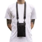 A person in a white t-shirt stands with a black crossbody pouch hanging from their neck. The pouch has a zippered compartment and a mesh pocket. The person has visible tattoos on both arms.