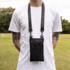 A person wearing a white t-shirt stands in a grassy field with trees in the background. They are wearing a black crossbody pouch with zippers, hanging from their neck by a strap, and have tattoos on their arms.