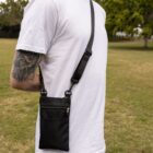 A person wearing a white T-shirt stands outside in a grassy area, carrying a black crossbody bag. The bag has a small front zipper pocket, and the persons arm has a visible tattoo. Trees are in the background.
