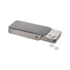 A metal tablet dispenser with a sliding mechanism. The dispenser is partially open, revealing a white tablet. Two additional white tablets are placed beside it on a white surface.