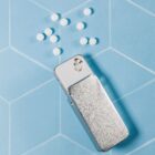 A metal pill dispenser is laying on a light blue, geometric-patterned surface with several small, round white pills scattered nearby.