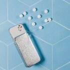 A metallic pill dispenser lies on a light blue geometric patterned surface. A few small, round white pills are scattered near the dispenser, with some emerging from its open top.