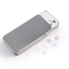 A small metallic rectangular tin with a sliding lid, partially open to reveal several white round mints inside. A few mints are scattered on the surface next to the tin.