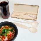 A bowl of noodle soup with shrimp and cilantro is next to a set of beige reusable cutlery, including a spoon, fork, and knife, stored in an open case. A glass of dark beverage is placed nearby on a white surface.