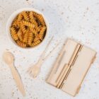 A bowl of rotini pasta sits on a terrazzo surface. Next to it is a beige reusable utensil set, including a spoon, fork, knife, chopsticks, and a straw cleaning brush, neatly arranged beside an open carrying case.