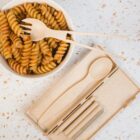 A bowl of spiral pasta with sauce, accompanied by a biodegradable spork. Beside it is an eco-friendly utensil set, including a spoon and reusable straws with a cleaning brush, all on a speckled countertop.