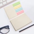 An open notebook on a white desk with colorful sticky tabs in yellow, green, blue, and pink. Nearby are a pair of glasses, a pencil, and part of a keyboard.