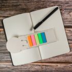 An open dotted notebook on a wooden surface, with a black pen placed diagonally across the pages. A set of brightly colored sticky notes with an attached ruler is on the left side of the notebook.