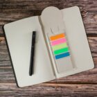 Open dotted notebook on a wooden surface with a black pen on the left and a bookmark with colorful sticky notes on the right. The sticky notes are orange, pink, green, yellow, blue, and teal.