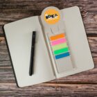 Open dotted notebook on a wooden surface with a black pen lying on top. There is also a bookmark containing colorful sticky tabs in pink, orange, green, blue, and yellow.
