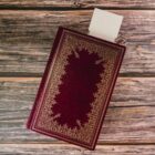 A closed book with an ornate gold floral border on a deep red cover lies open on a wooden surface. A blank paper protrudes from the top of the book, suggesting its used as a bookmark.