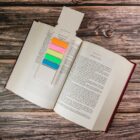 An open book on a wooden surface with colorful sticky tabs on top. The tabs are pink, orange, yellow, green, and blue, attached to a piece of cardboard.