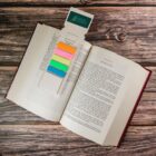 An open book on a wooden surface with a bookmark holding colorful sticky tabs. The pages are marked with text, and the bookmark has an attached card that says Green Spaces with a tree logo.