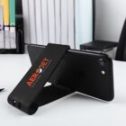 A smartphone rests horizontally on a black phone stand branded with AeroJet in red text. The stand is placed on a white desk with blurred office supplies and furniture in the background.
