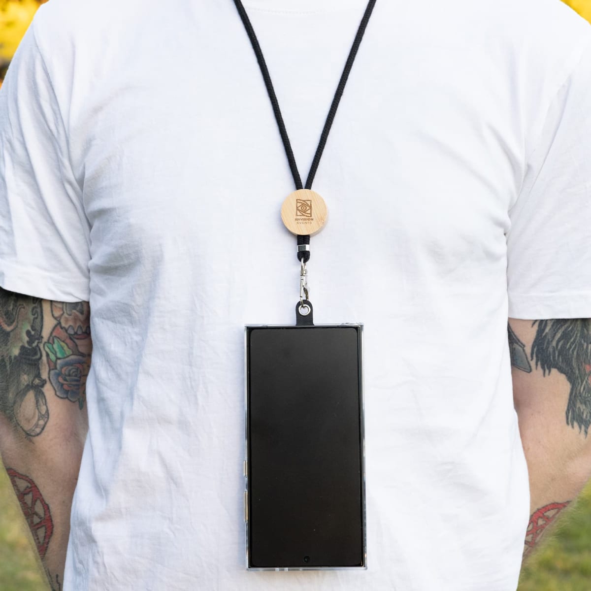 A person wearing a plain white t-shirt is shown with a smartphone hanging from a black lanyard around their neck. The persons arms have colorful tattoos, and the lanyard has a wooden attachment. The background is blurred greenery.
