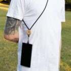 A person wearing a white t-shirt with tattoos on their arm is standing outside on grass. They have a smartphone hanging from a black lanyard around their neck. The phone is positioned at their side.