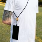 A person wearing a white t-shirt and showing arm tattoos is standing on grass. They have a smartphone attached to a wooden holder hanging from their neck with a black strap.