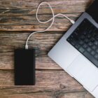 A silver laptop connected to a black external hard drive with a white cable rests on a textured wooden surface. The laptops keyboard and trackpad are partially visible, while the hard drive lies beside it.
