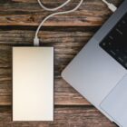 A silver external hard drive is connected to a laptop with a white cable. The laptop is partially visible, showing a corner of the keyboard. They are placed on a rustic wooden surface with a textured, weathered look.