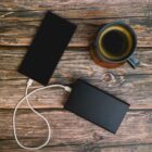 A smartphone is connected to a power bank with a white cable on a wooden surface. A cup of coffee sits nearby.