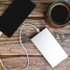 A smartphone connected to a power bank with a white cable is on a wooden table. A cup of black coffee sits nearby on a coaster.