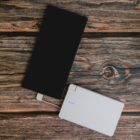 A smartphone connected to a white power bank with a short cable. Both devices are placed on a wooden surface with visible grain and knots.