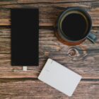 A smartphone charging with a white power bank, next to a cup of black coffee, all placed on a wooden surface.