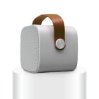 A white portable speaker with a brown leather strap sits on a minimalist white pedestal. The speaker has a rounded square shape and features a textured front grill.
