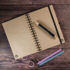 Open spiral-bound notebook with lined pages on a wooden table, featuring a black pen resting on it. Two markers, one pink and one purple, lie below the notebook.