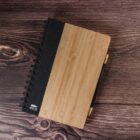 A top-down view of a spiral-bound notebook with a wooden cover on a dark wooden table. A pen is securely attached to the notebooks cover. The texture of both the notebook and the table is prominently visible.