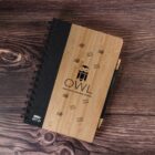 A top-down view of a spiral-bound notebook with a wooden cover on a wooden surface. The cover features a cartoon owl and the text OWL Scholastic Tutorial Center. A pen is attached to the notebook.
