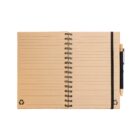 An open spiral notebook with brown recycled paper and lined pages. A black elastic band holds a black pen on the right side. The pages have recycling symbols at the bottom corners.