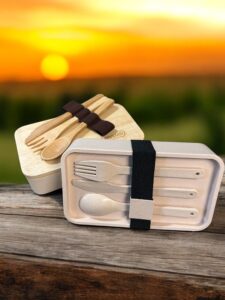 Two eco-friendly promotional lunchboxes with wooden covers and utensils are secured by black elastic bands. One is open, displaying a fork, spoon, and knife. Perfect for Back to School 2025, they are placed on a wooden surface with a vibrant sunset in the background.